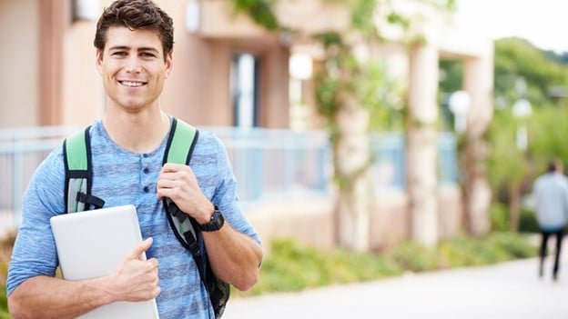 a boy carrying a laptop in his hand and a black and green backpack in his shoulder.